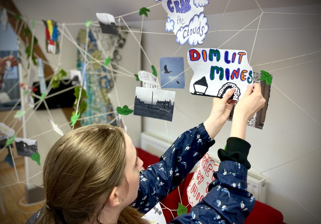 A young woman pins a hand-drawn picture to a piece of string in an installation. The picture reads 'Dim lit mines'.
