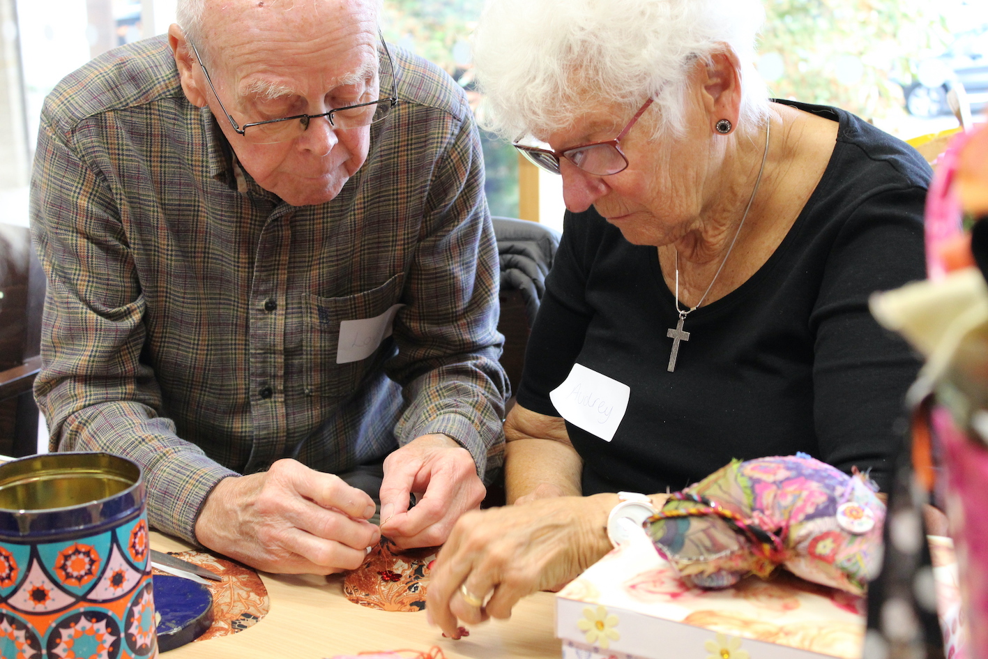 Two old people sitting at table sewing fabric in a room