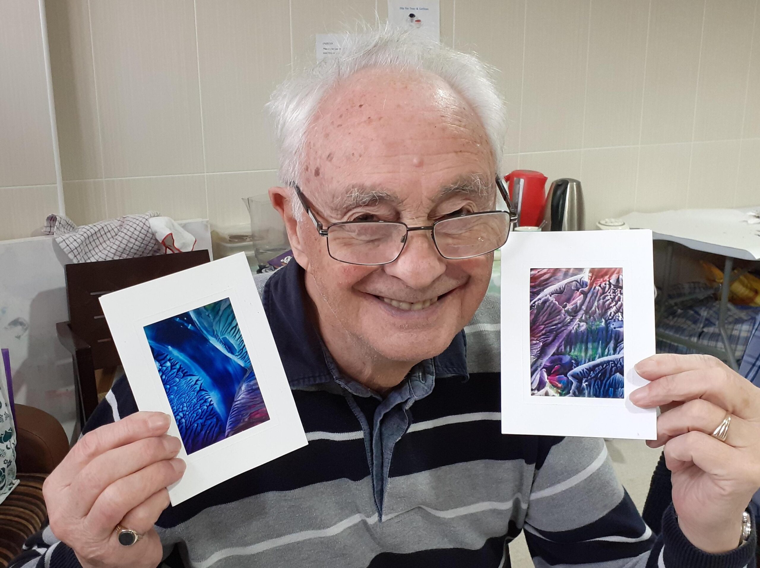 Old man sitting holding up two paintings smiling in a room