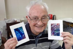 Old man sitting holding up two paintings smiling in a room