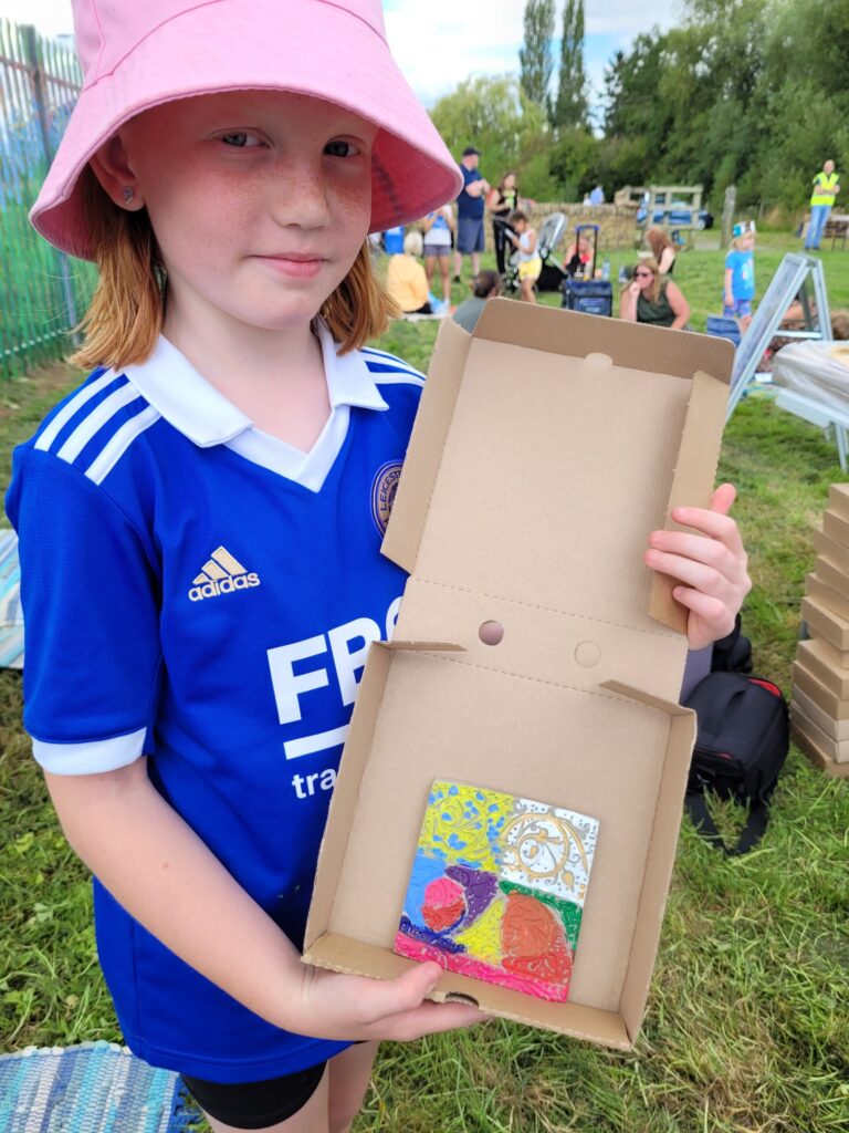 Girl in blue shirt and pink hat shows an open box with her artwork in.